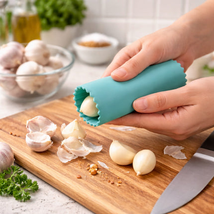 Person using a teal garlic peeler on a wooden cutting board with garlic and herbs in the background.