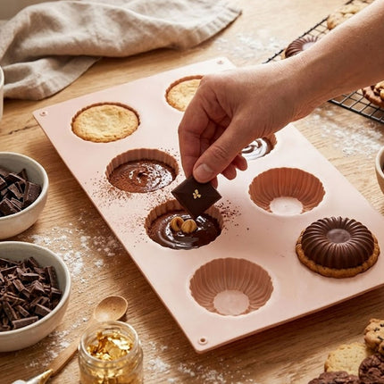 Person filling a silicone mold with chocolate and nuts on a wooden table.