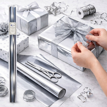 Person wrapping a gift with silver wrapping paper and decorative items on a marble surface