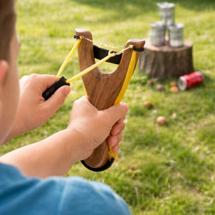 Child holding a wooden slingshot in an outdoor setting with grass and a tree stump.