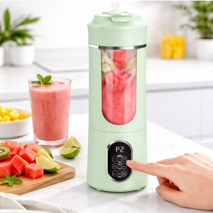 Green portable blender with a smoothie inside, surrounded by fruits and a glass of juice on a kitchen counter.