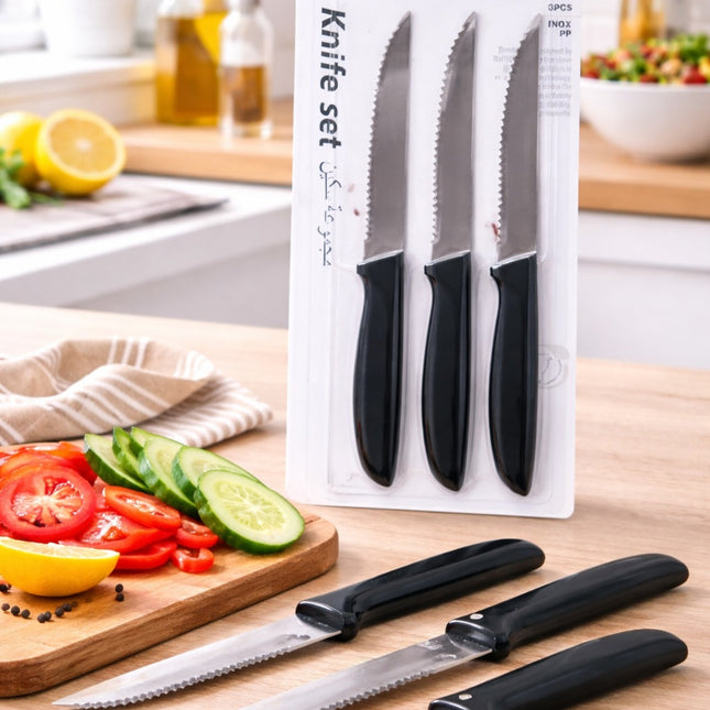 Set of three knives with black handles on a wooden cutting board with sliced vegetables and a kitchen background.