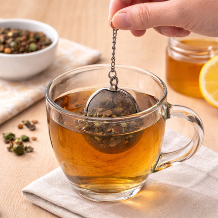 Clear glass mug with tea and a metal tea ball, surrounded by tea leaves and a bowl on a wooden surface.
