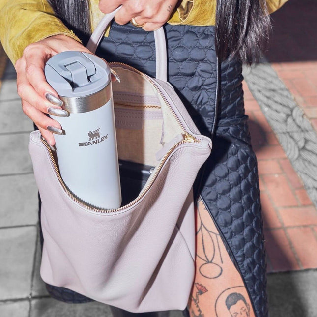 A woman holds a Stanley travel tumbler in the side pocket of a light pink leather shoulder bag. The bag is shown in a close-up outdoor shot, with the woman wearing a quilted navy and mustard yellow jacket.