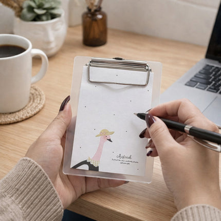 Person holding a clipboard with a design of an ostrich wearing a hat, sitting at a desk with a laptop and coffee cup.
