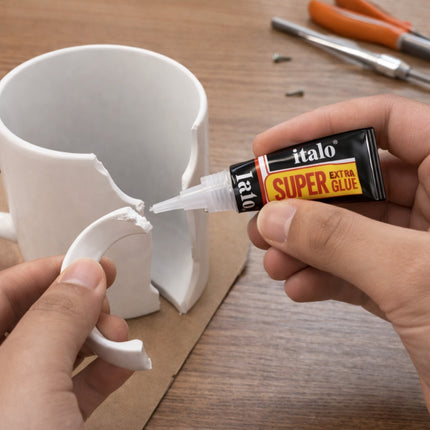 Person using italo Super Extra Glue to repair a broken white mug on a wooden surface.