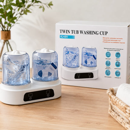 A twin tub washing cup set with two clear blue water reservoirs on a white base, shown next to its retail box on a wooden tabletop with a vase and basket as props.