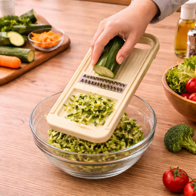 Person using a vegetable chopper to shred a cucumber over a glass bowl on a wooden table with vegetables around.