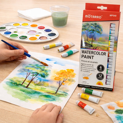 Person painting a landscape with watercolor paints on a wooden table, Motarro watercolor paint set visible.